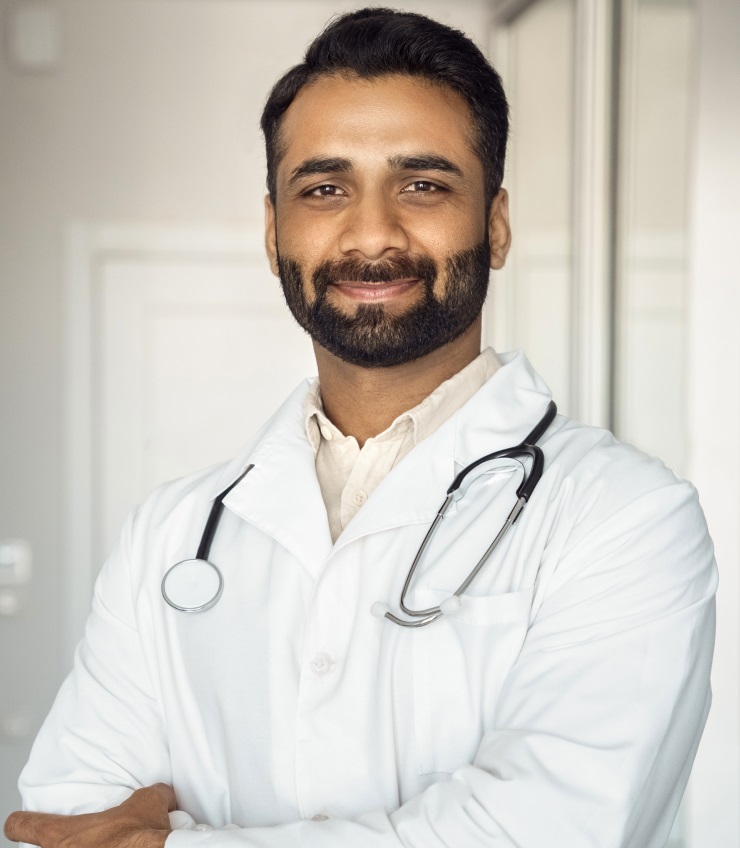 Vertical Portrait Of Smiling Young Indian Male Doctor In Medical Robe Uniform Standing Arms Crossed In Hospital Office